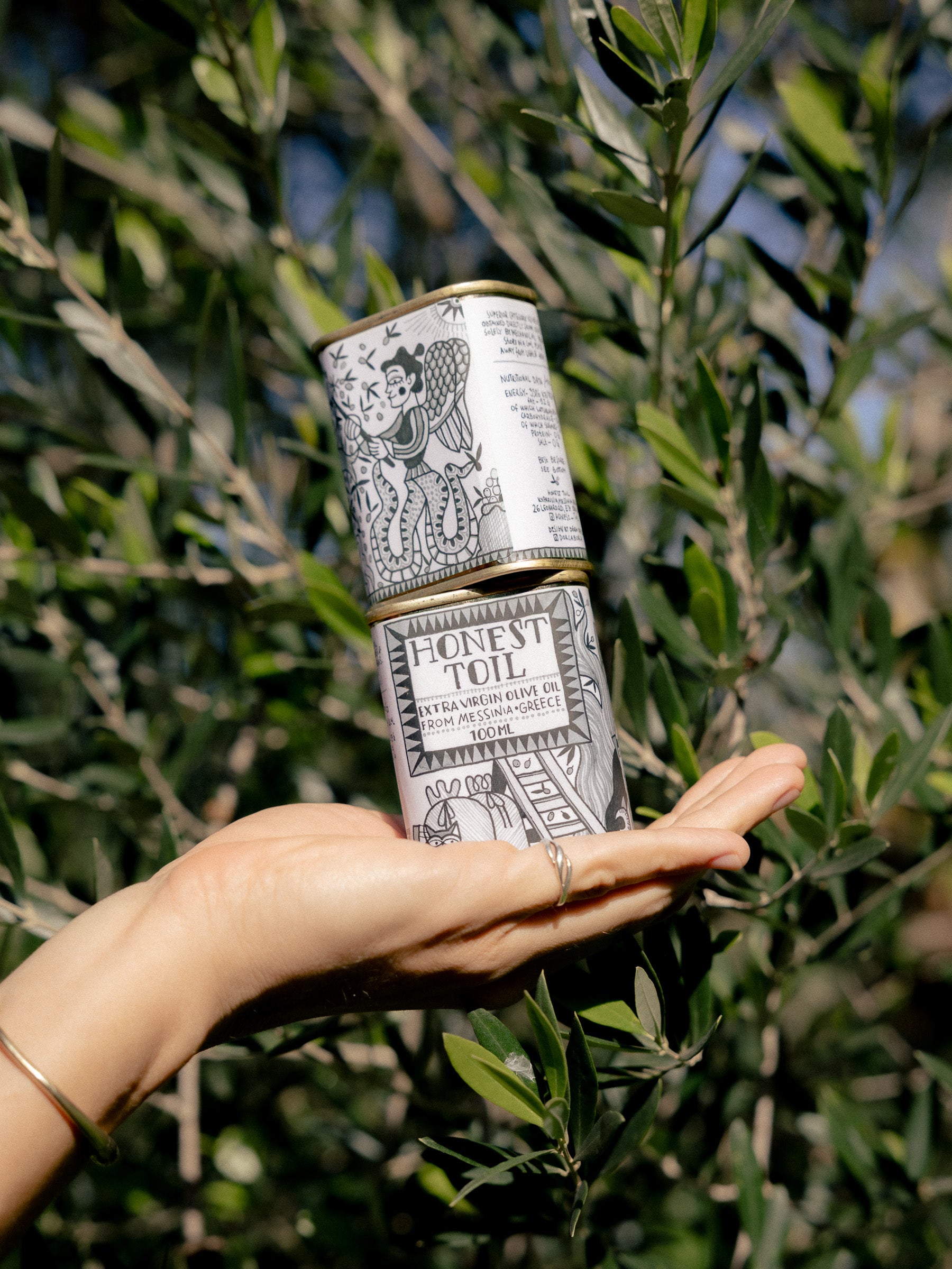 A hand holding two stacked Honest Toil 100ml tins in front of olive tree branches.