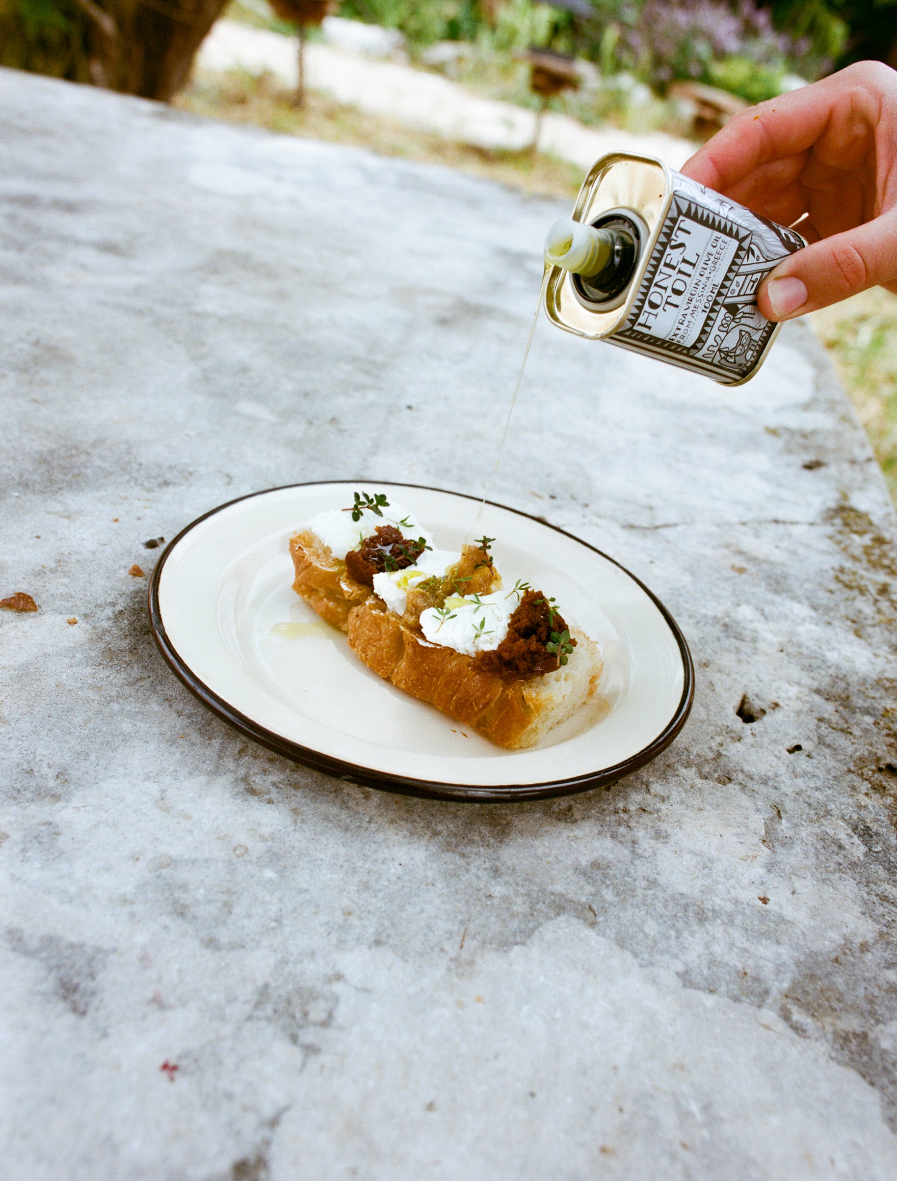 Olive oil being poured from a Honest Toil 100ml tin onto bruschetta with ricotta and fig on a plate.